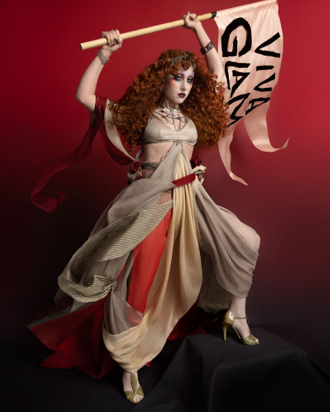 Chappell Roan appears in a MAC Viva Glam ad wearing a flowing costume and holding a flag. Her hair is styled in curls...